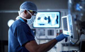 A male doctor dressed in blue scrubs, a white surgical mask, blue rubber gloves, and glasses looking at a monitor