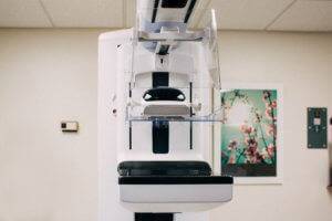 A white and black mammogram machine in a white room with a poster of pink flowers hanging on the wall